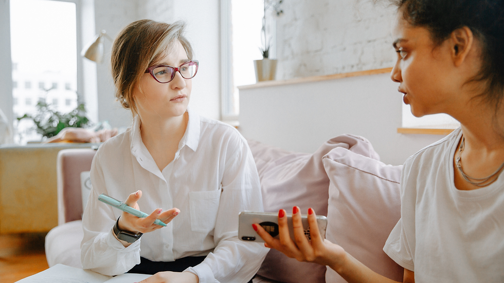 Two women talk on a couch in a bright living room, one holding a pen and notebook, the other a phone.