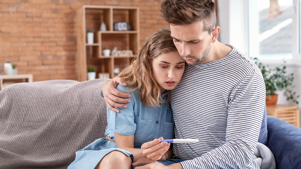 A couple on couch, woman in blue dress, man in striped shirt, holding a pregnancy test. Concerned expressions, brick wall background.