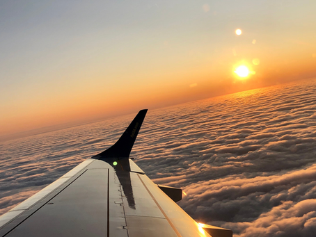 Airplane wing over fluffy clouds at sunset. Orange sky and sun create a serene mood. The wing reflects warm light, enhancing the scene.