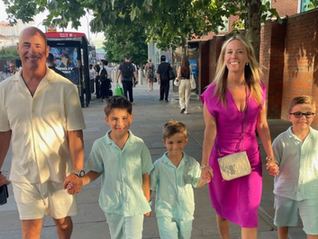 Family walking on a sidewalk on a sunny day. Adults in white and pink attire, kids in light blue outfits. Tree-lined street in the background.