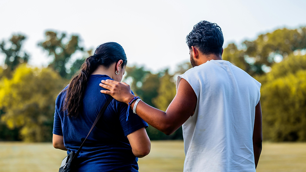 Man in white shirt with his arm around a woman in a blue shirt, walking in a sunny park with trees. Mood is supportive and caring.