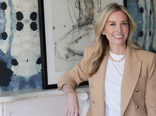 Smiling woman in tan blazer and white shirt stands by a marble surface. Abstract blue and black art decorates the wall behind her.