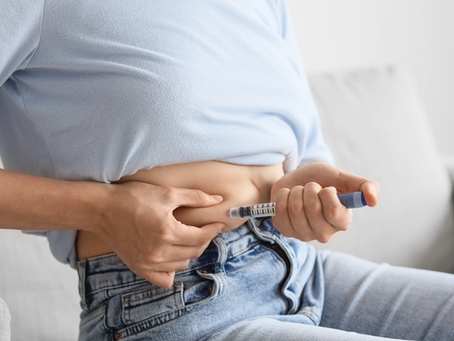 Person in a light blue shirt self-administering an injection in the abdomen, sitting on a gray couch. Focused, calm setting.