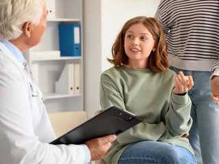 A doctor talks to a seated woman in a green sweater, while another person stands nearby. Office setting with shelves in the background.