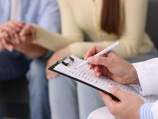 Doctor writes on clipboard while a couple holds hands in an office. The mood is supportive and reassuring.