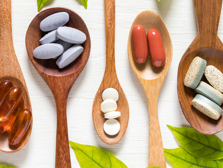 Assorted pills and capsules in wooden spoons on a white surface, framed by green leaves, arranged neatly.