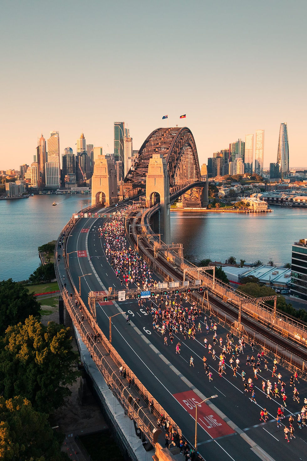 Aerial view of marathon runners crossing the Sydney Harbour Bridge at sunrise, with the city skyline behind.