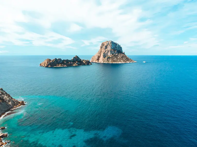 Rugged islets near Ibiza, including a towering rock stack, surrounded by vivid blue water and light clouds
