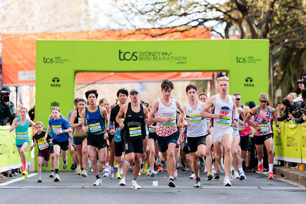 Runners of all ages surge from the start line beneath the green TCS Sydney Mini Marathon arch on a tree-lined city street.