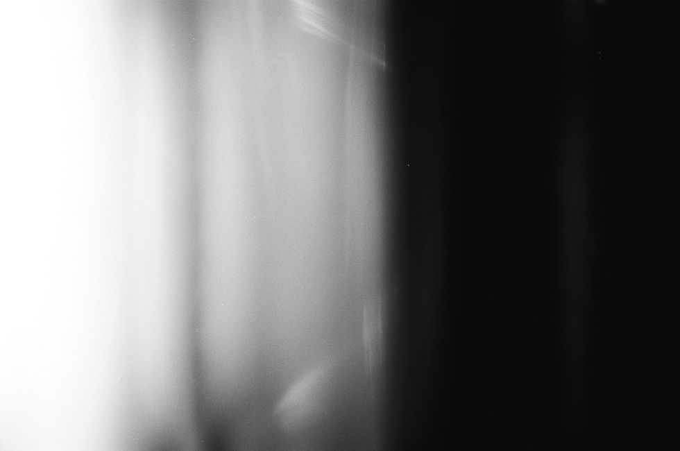 Ein abstraktes Schwarz-Weiß-Foto, auf dem auf der linken Seite schlierenhaft ein Vorhang vor einem Fenster zu sehen ist. Er ist hell beleuchtet, die zweite rechte Bildhälfte ist ganz dunkel.