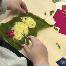 a photograph of a person's hands and a wet-felted piece. the right hand is holding a small pair of scissors while the left hand is holding the corner of the felted piece. The felted piece has sa dark green background and has a baby chick in yellow with an orange beak and a black eye in the foreground. The chick has bright purple feet.