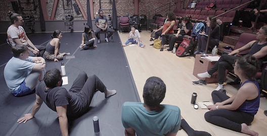 Artists at ‘Signals from the West’ Conversation are in discussion, sitting in a large circle on the floor and in seats in a performance space with their personal belongings.