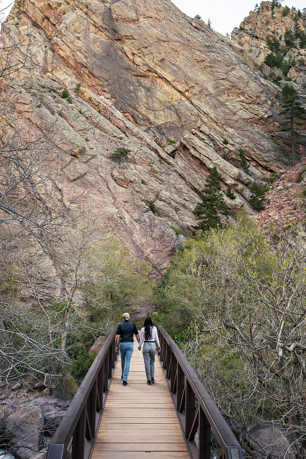 Engaged couple walks hand-in-hand across a wooden footbridge toward towering sandstone canyon walls at Eldorado Canyon State Park near Boulder, Colorado