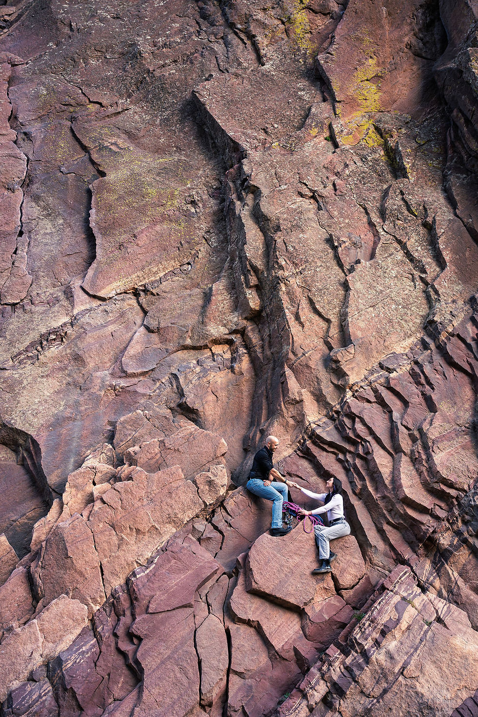Aerial view of an engaged couple sitting on a massive red sandstone cliff face at Eldorado Canyon State Park near Boulder Colorado, holding hands with climbing rope beside them