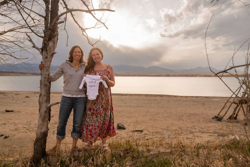 two women standing next to each other holding a baby onesie that says we are a family