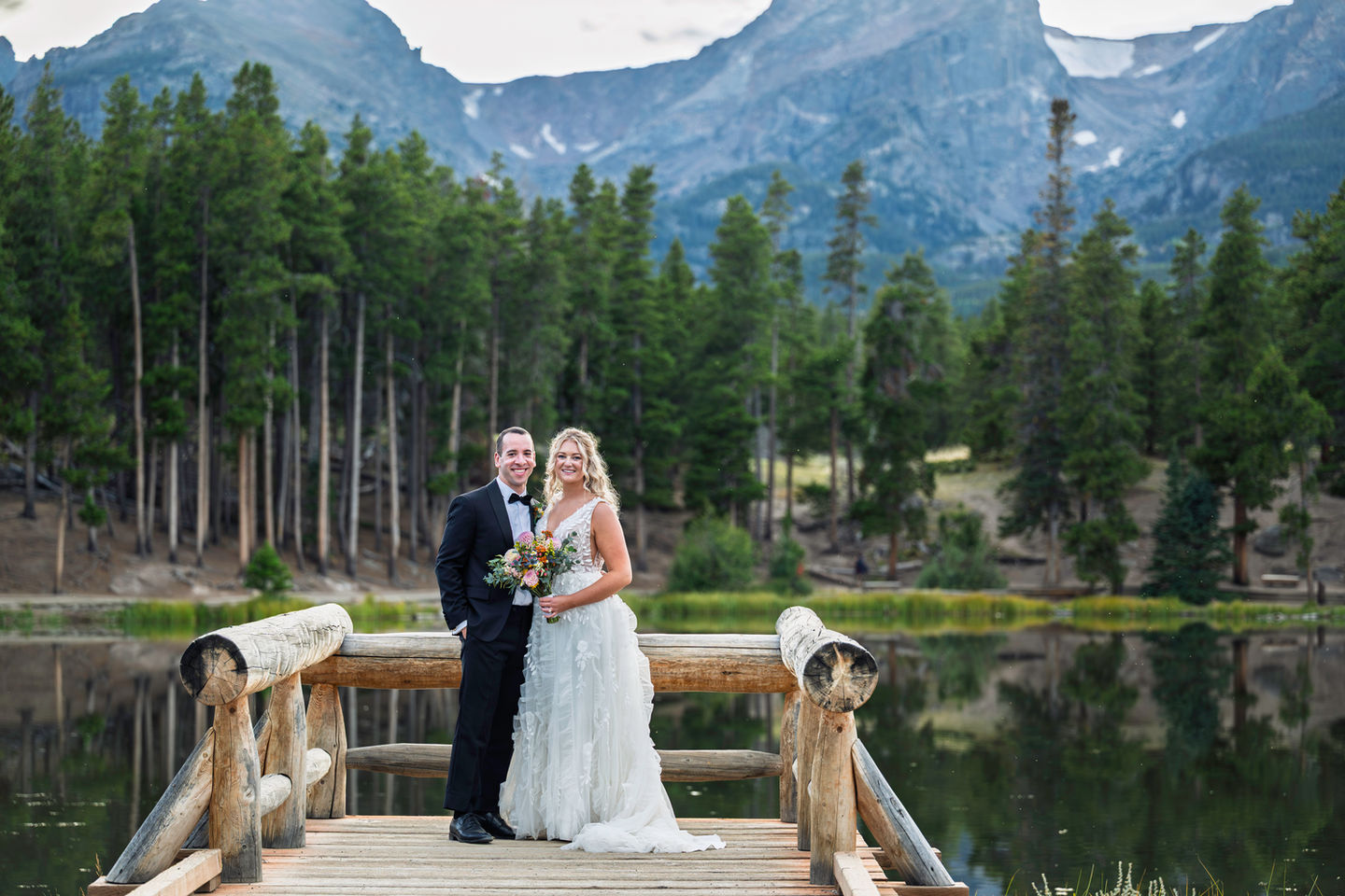 Bride and groom on a wooden bridge, mountainous backdrop.