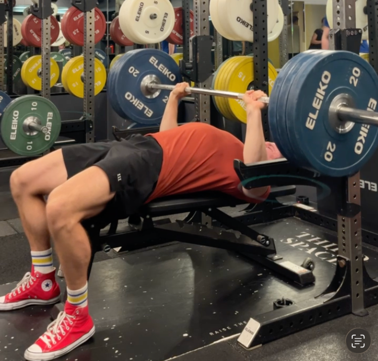 Man bench presses a barbell loaded with Eleiko plates in a gym rack, wearing a red shirt and red Converse.
