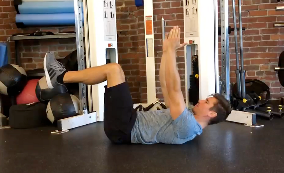 Man doing a crunch on gym floor, knees bent and arms raised, with exercise equipment and a brick wall behind him.