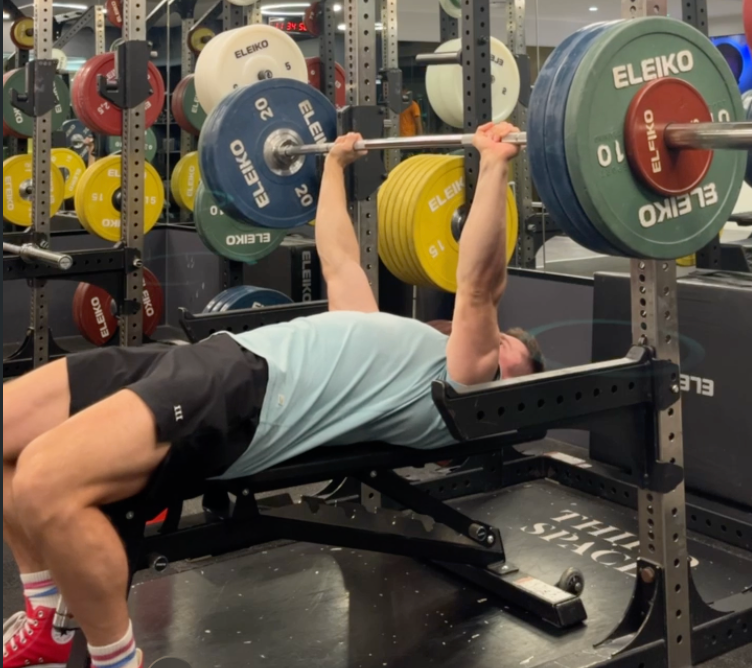 A person bench presses weights in a gym. The rack is filled with colorful weight plates marked "ELEIKO." The setting is modern and bright.
