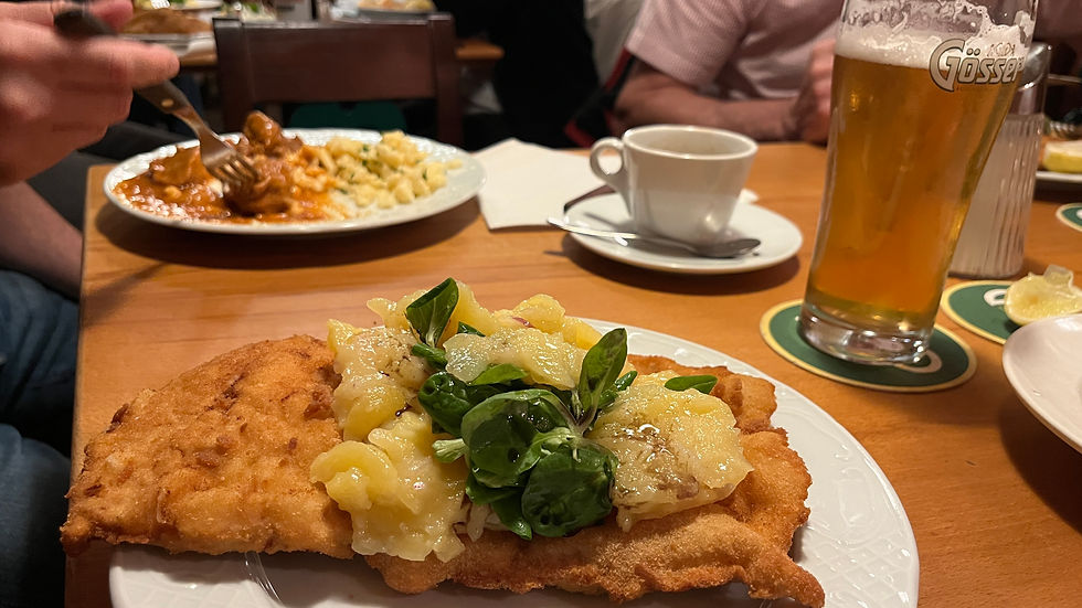 Plate with schnitzel, potato salad, and greens on a wooden table. Nearby, a hand holds a fork, a beer glass, and a coffee cup are visible.