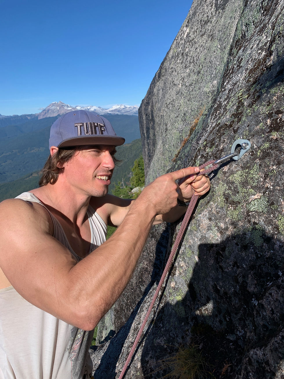Climber in a TURF cap clips rope to an anchor on a sunlit granite cliff, with snowy mountains and blue sky behind.