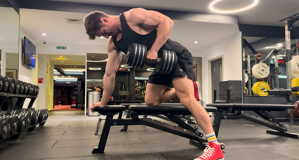 Man lifting a heavy dumbbell on a gym bench, wearing a black tank top and red sneakers. Bright gym with mirrors and equipment.