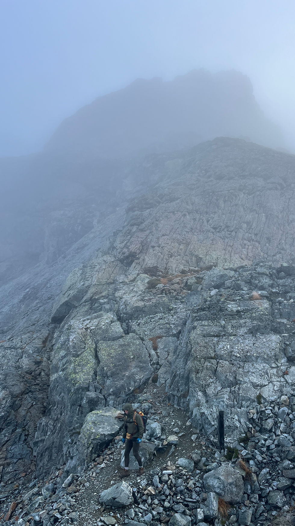 Hiker with backpack climbs a rocky, fog-shrouded mountain trail, surrounded by steep gray cliffs and mist.