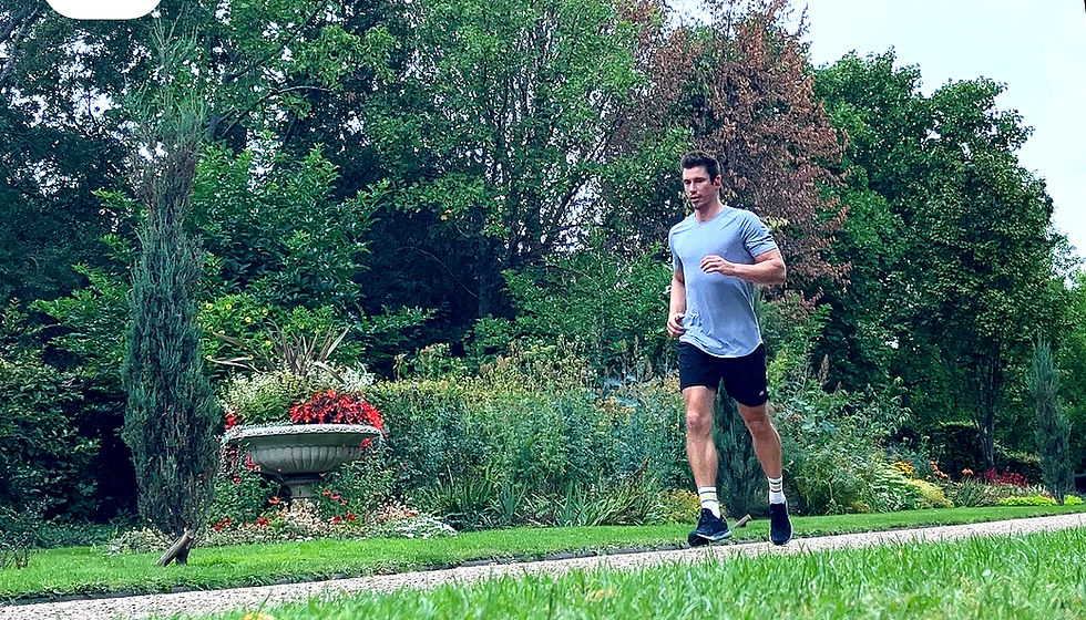 Man jogging in a park with lush green trees and colorful flowers. He's wearing a gray shirt and black shorts, exuding a focused mood.
