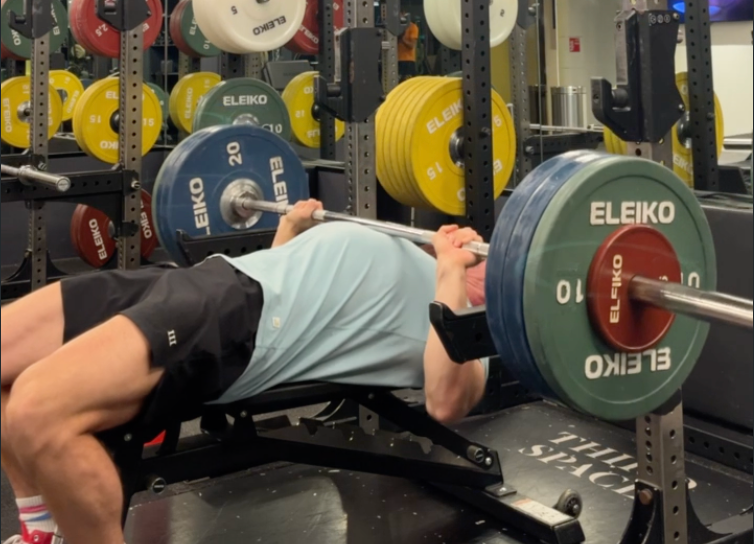 Man in a blue shirt bench pressing heavy weights in a gym. Colorful plates labeled "Eleiko" are visible. Focused and intense setting.