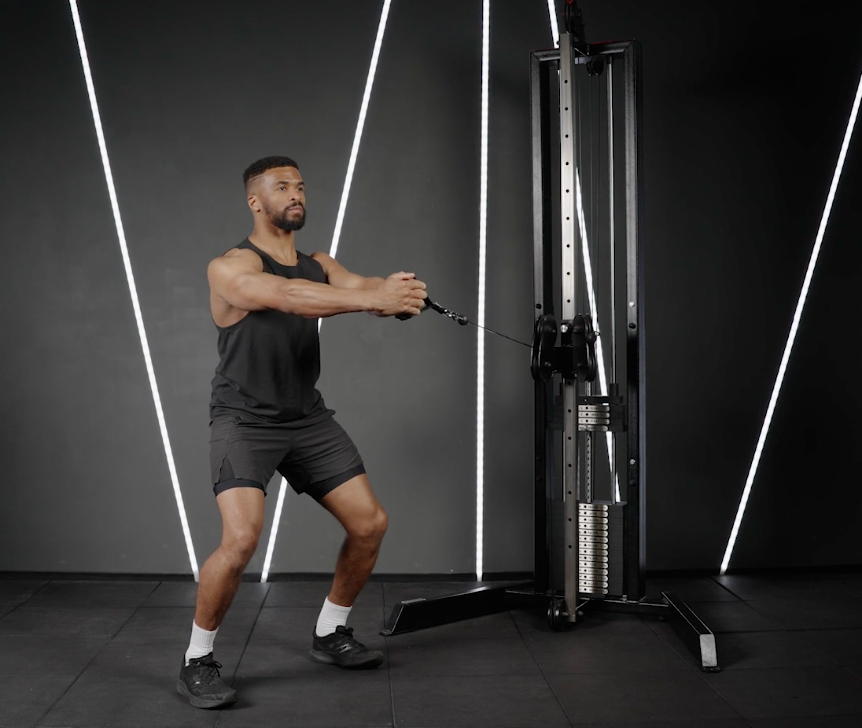 Man in black workout clothes performs a cable pull on a gym machine in a dark studio with bright white diagonal light strips.