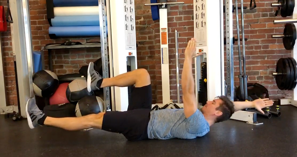 Man doing a core exercise on a gym floor, one arm raised, legs bent and lifted, with medicine balls and cable machines behind.