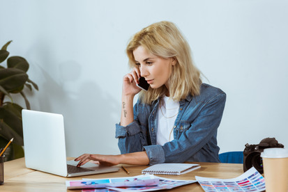 Young female entrepreneur sitting down at her desk, talking on the phone, and working on her laptop