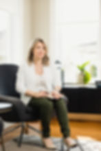 Laurence Ste-Marie in a white cardigan sits on a chair, holding a notebook and pen. Bright room with large windows and a plant in the background.