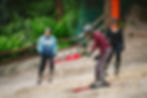 A man in a red shirt and grey trousers tries a practise run on skis down an outdoor all-weather ski slope. There are other students in the background also involved in the ski lesson.