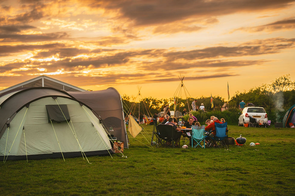 The sun sets over a camping field in Somerset, with a family gathered around a camp table next to their tent.