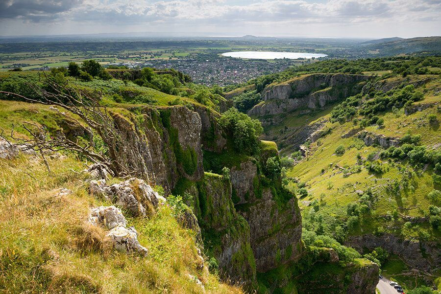 View of Cheddar Gorge from one side, looking towards Cheddar Reservoir. Home to nature and adventure activities, rugged cliffs drop down out of sight into the gorge, while the rolling Mendip Hills are covered in lush green trees and grassland.