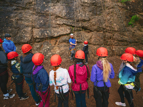A group of children wearing rock climbing harnesses and helmets stand in a semi-circle, facing a rock face and the instructor standing in front of them. They are listening to his instructions while on a Rock Climbing session.