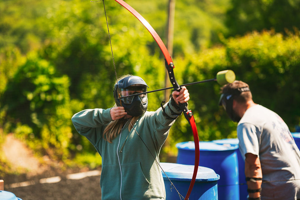 A person with long hair wears a black paintball-style mask and is pulling back the string on a bow they have loaded with a foam-tipped safety arrow.