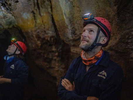 Two people in a cave, wearing helmets and headtorches, look upwards while gently smiling.