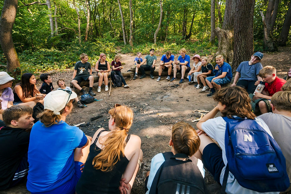 A large group of young people on an outdoor learning trip sit in a circle in a woodland, chatting and laughing. There are a few outdoor instructors in blue shirts sat with them.
