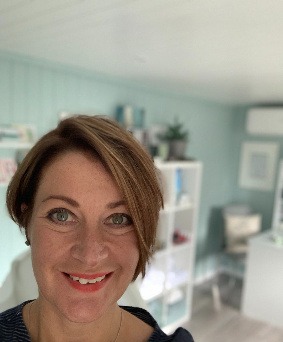 Woman smiling indoors with short brown hair. Light blue walls and white shelves in background. Bright, cheerful atmosphere.