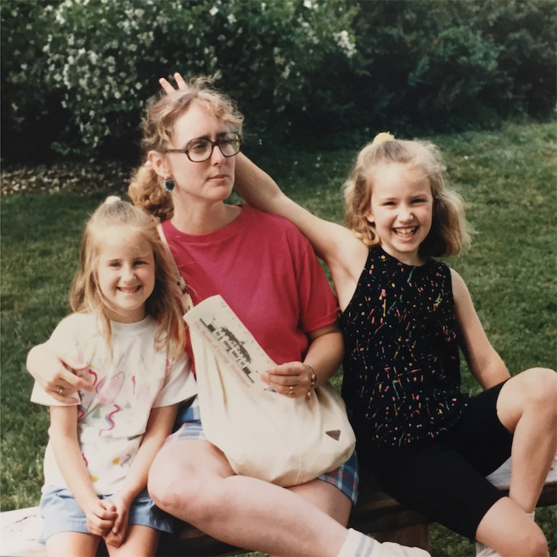 Family photo from the late 80s, white woman in her 30s with big tortoiseshell glasses and permed honey blonde hair, with two young girls on other side of her. They are sitting on a bench and the older one on the right is giving her bunny ears.