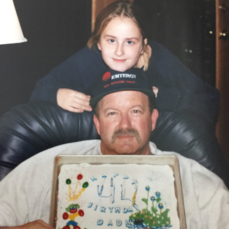 Father and daughter, daughter resting her head on her dad's while he sits in an armchair showing his birthday cake.