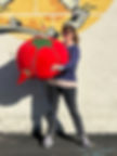 Woman holding a gigantic 2 foot wide tomato pincushion she has made by hand. It also has some giant pins and a needle in it.