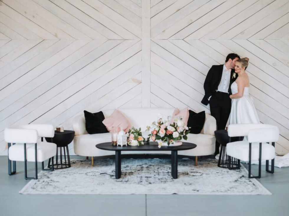 Bride and groom stand beside a modern sofa with floral arrangement in a chic, white-paneled room. Elegant and romantic atmosphere.
