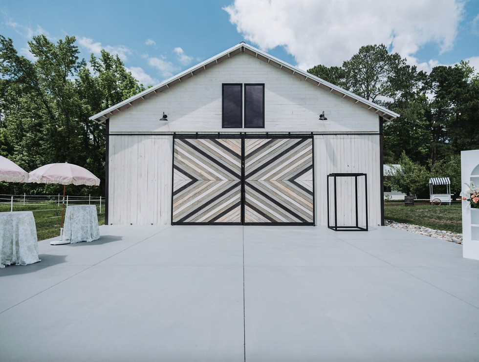 Barn with geometric doors, surrounded by trees. Two tables with umbrellas and a floral stand on a sunny, clear day. Bright and serene mood.