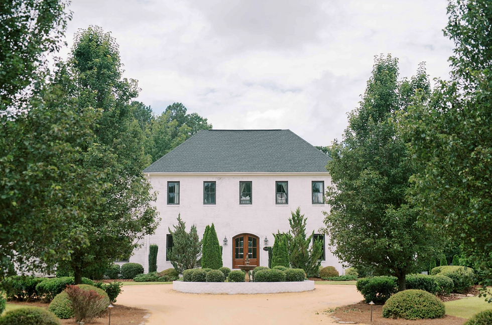 Large white house with dark roof, surrounded by lush greenery and trees. Overcast sky and tranquil atmosphere. No visible text.