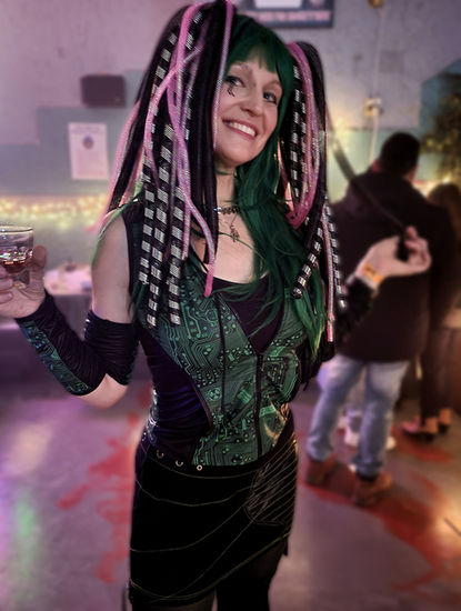 A woman with green hair and a green and black motherboard print outfit wears black and pink cyberlocks. The woman smiles with her head cocked, her hands raised with arms bent at the elbows, and a cocktail in her right hand.