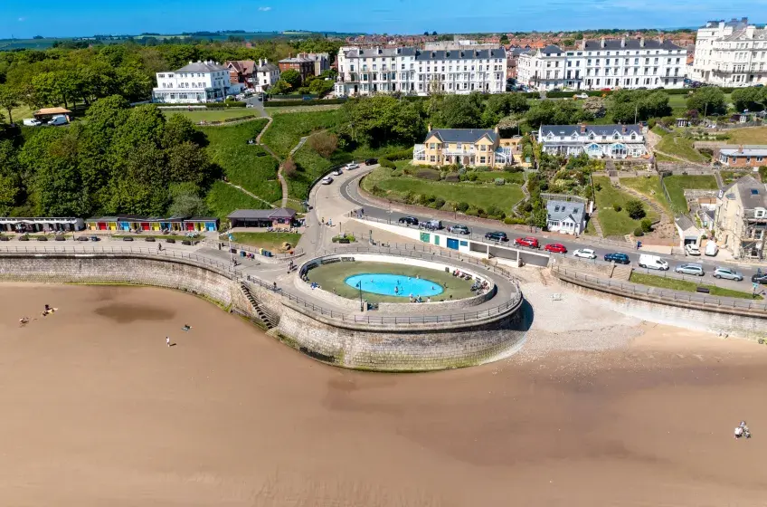 An aerial view of Filey Beach near Bungalow Farm in Buckton, showing the sandy shore, tidal pool, promenade, colourful beach huts and coastal town backed by green hills.