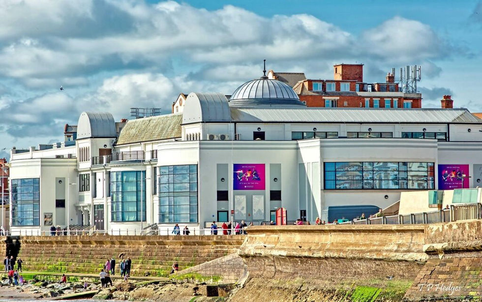 The modern white‑fronted Bridlington Spa near Bungalow Farm in Buckton, with large glass windows, colourful show posters and people walking along the seafront promenade on a bright day.